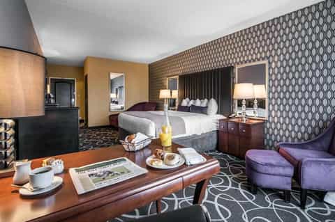 Luxury hotel suite with king bed, sitting area, patterned wallpaper, wooden desk, and purple accent chairs