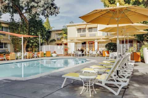Resort pool with white lounge chairs, yellow cushions, large yellow umbrellas, tropical landscaping, and residential buildings