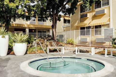 Circular hot tub on concrete patio with white railings, large tree, and yellow-striped awning building