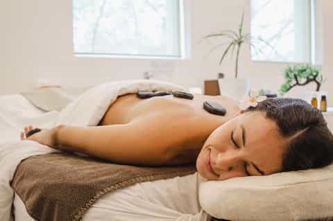 Woman receiving hot stone spa massage treatment on white bed in serene wellness room with windows.