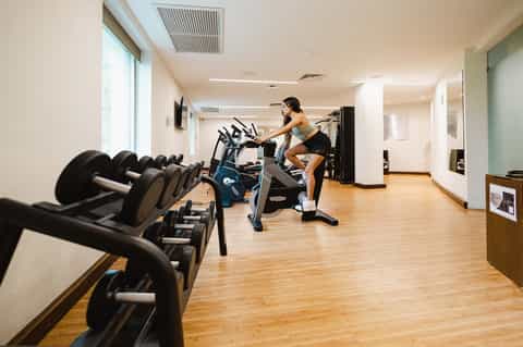 Woman on stationary bike in modern gym with dumbbells, bright windows, and wooden flooring