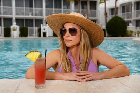 Woman in purple swimsuit enjoying cocktail at resort pool wearing sun hat and sunglasses