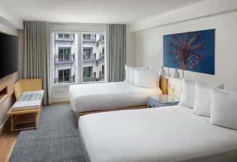 Hotel room with two queen beds, wooden headboards, modern decor, and city views through tall windows