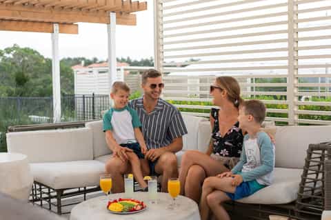 Family relaxing on modern deck with beige sofa, tropical views, wooden pergola, and slatted privacy screens