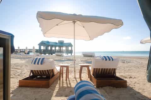 Private beach cabana with two striped loungers under cream umbrella, ocean view, and resort pavilion visible
