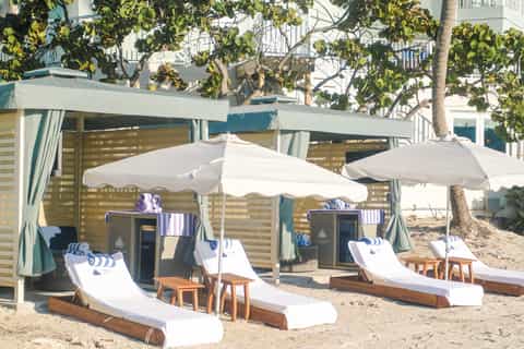 Beach cabanas with white umbrellas and loungers on sand, covered seating areas with tropical plants