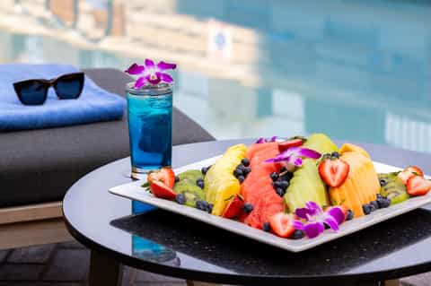 Poolside table with colorful fruit plate, blue cocktail, pink orchid, and city skyline backdrop