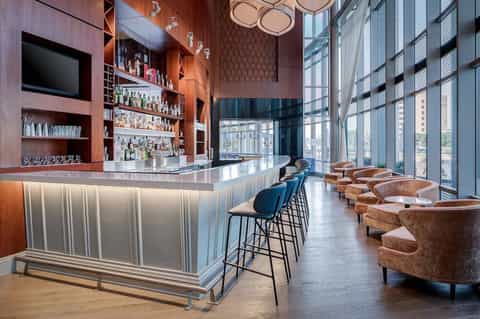 Modern bar lounge with wooden shelving, pendant lighting, cognac leather seating, and floor-to-ceiling windows