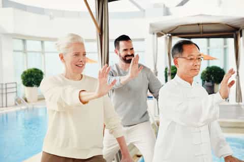 Three people doing tai chi by an indoor pool with large windows and palm plants