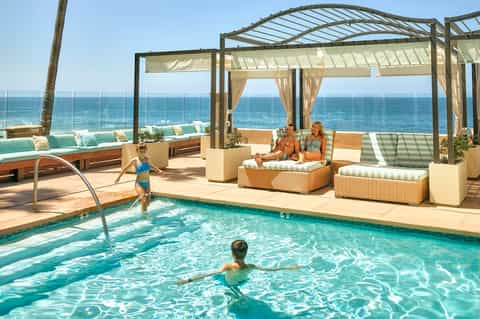 Beachfront resort pool with pergola shade structures, lounge seating, ocean view, and family enjoying water