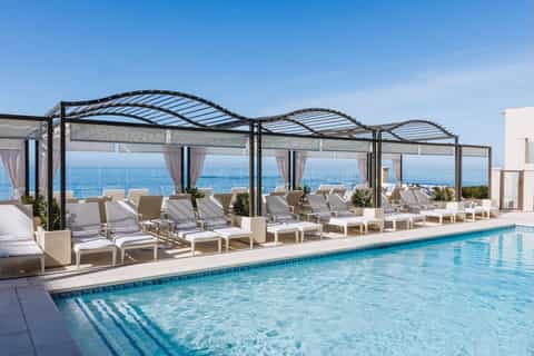 Luxury rooftop pool with ocean horizon, white cabanas, pergolas with shade, and beige lounge seating