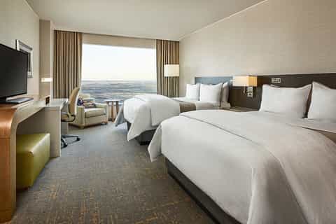 Modern hotel room with two beds, expansive window view, flat-screen TV, and neutral décor