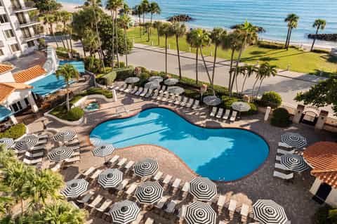 Aerial view of curved resort pool with striped umbrellas, ocean, and palm trees
