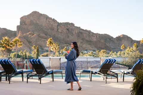 Woman on resort sundeck with lounge chairs, mountain backdrop, palm trees, and pool view