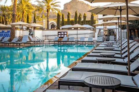 Resort pool with turquoise water, black lounge chairs, tan umbrellas, mountains, and desert landscaping