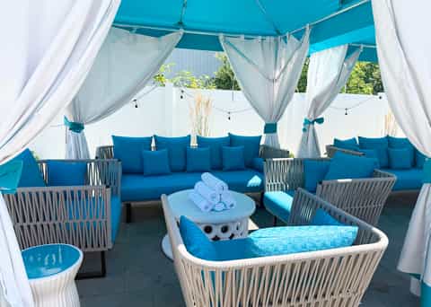 Luxury cabana lounge with blue cushions, white curtains, rolled towels, and shaded seating