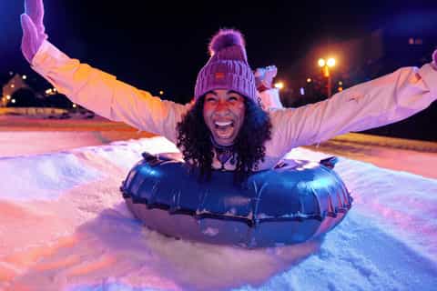 Person tubing on snow at night under colorful lights with winter lodge backdrop
