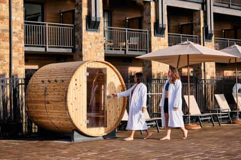 Guests in white robes viewing barrel sauna on wooden deck with hotel buildings, umbrellas, and loungers