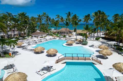 Beachfront resort pool complex with turquoise water, palm trees, tiki umbrellas, and ocean view