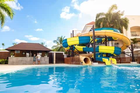 Resort pool with yellow and blue waterslide, palm trees, and tropical resort buildings