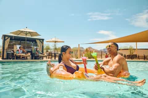 Couple enjoying pool floats and colorful drinks at resort pool with cabanas and shade structures