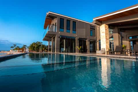 Modern waterfront home with infinity pool overlooking beach and palm trees at dusk