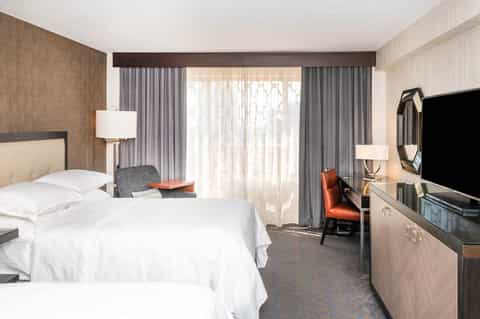 Hotel bedroom with queen bed, modern furnishings, work desk, balcony doors with curtains, and flat-screen TV