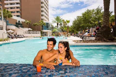 Couple swimming in resort pool with palm trees, high-rise hotel building, and tropical landscaping