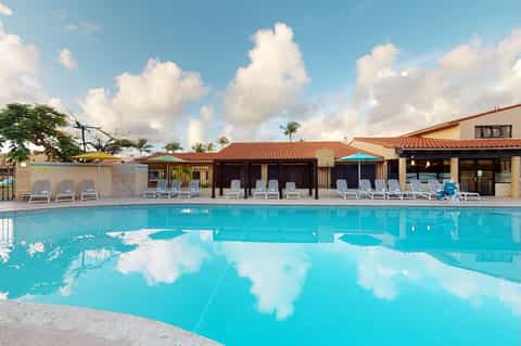 Resort pool reflecting sky with cabanas, lounge chairs, palm trees, and Spanish-style buildings