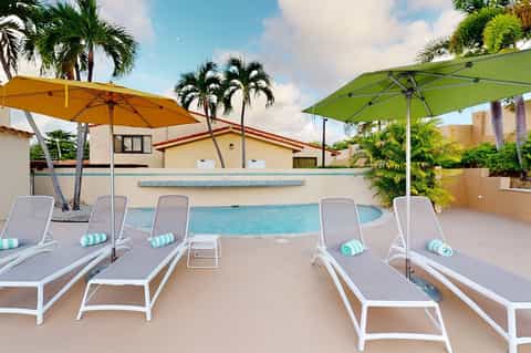 Resort pool with white lounge chairs, colorful umbrellas, palm trees, and tropical building architecture