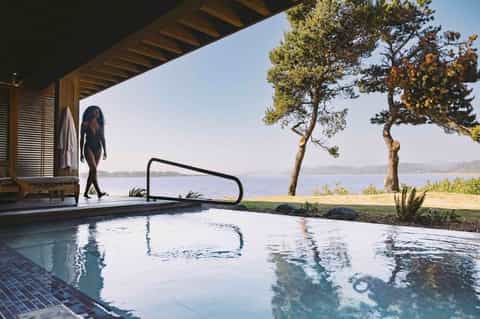 Woman in swimwear approaching natural hot spring pool overlooking lake and forested landscape