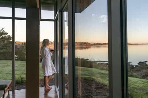 Woman in white robe viewing calm lake from modern glass doors at sunrise