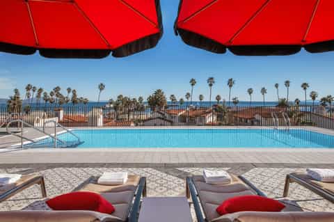 Beachfront pool under red umbrellas with palm trees, ocean, and residential buildings in background