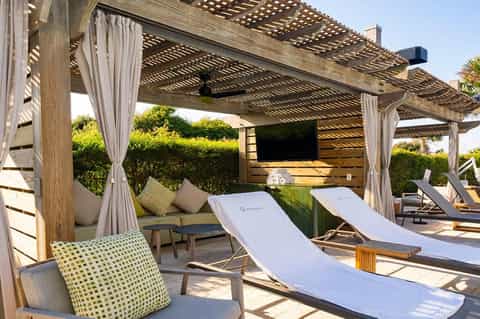 Luxury cabana with wooden pergola, lounge chairs, flowing curtains, and integrated TV screen