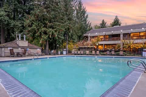 Resort pool at twilight surrounded by evergreen trees with lit lodge building and fire pit area