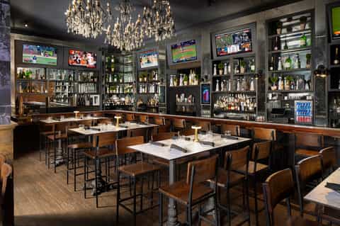 Upscale restaurant interior with crystal chandeliers, extensive liquor display, TV screens, and wood furnishings