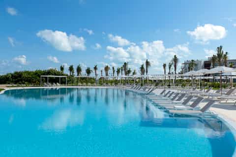 Large resort swimming pool with crystal-clear turquoise water, lounge chairs, umbrellas, and palm tree landscaping