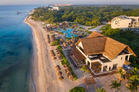 Aerial view of beachfront resort with thatched-roof structures, pools, and pristine white sand beach