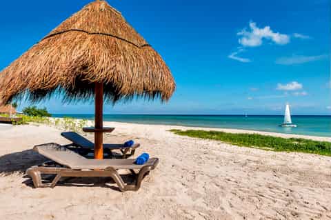 Tropical beach with thatched umbrella, lounge chair, sailboat, and clear turquoise water