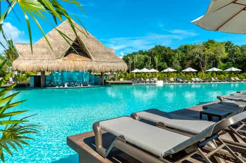 Luxury resort pool with thatched-roof bar, turquoise water, white umbrellas, lounge chairs, and jungle backdrop