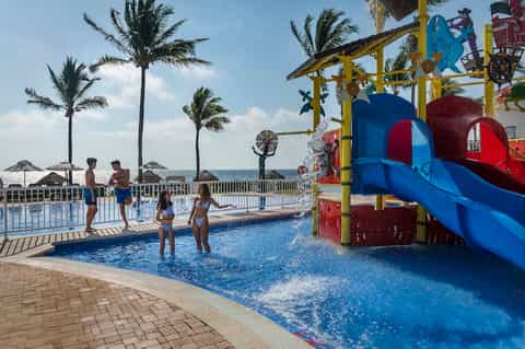 Resort pool area with colorful children's water slide, palm trees, beach umbrellas, and ocean view