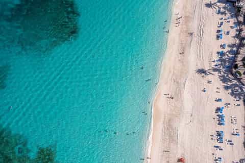 Aerial beach view with turquoise water, white sand, palm tree shadows, and blue lounge chairs
