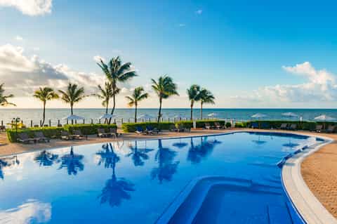 Beachfront resort pool with palm tree reflections, lounge chairs, beach access, and ocean horizon