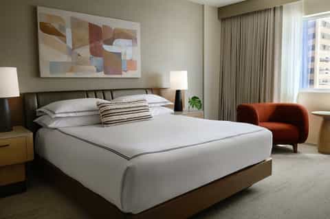 Modern hotel bedroom with king bed, contemporary artwork, rust-colored chair, and city views
