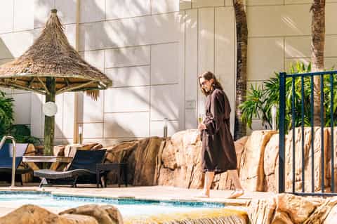 Resort pool area with modern furniture, thatched umbrella, hanging light, stone water feature, and palm trees