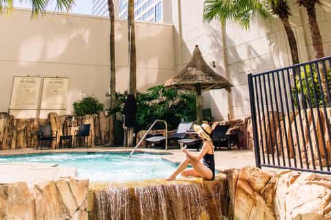 Indoor tropical pool area with palm trees, thatched umbrella, stone walls, and modern hotel architecture