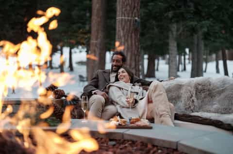 Couple by firepit at resort during evening with fire, blankets, drinks, and forest backdrop