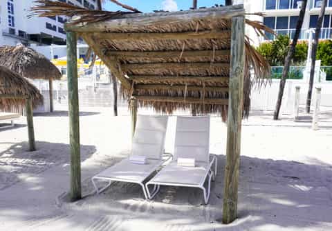Wooden tiki-style shade structure with two white lounge chairs on sandy beach area