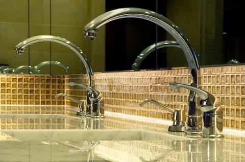 Bathroom vanity with dual chrome faucets, beige tile backsplash, and illuminated mirror in contemporary design