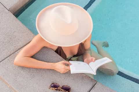 Person in pool lounging on float while reading magazine wearing sun hat and sunglasses